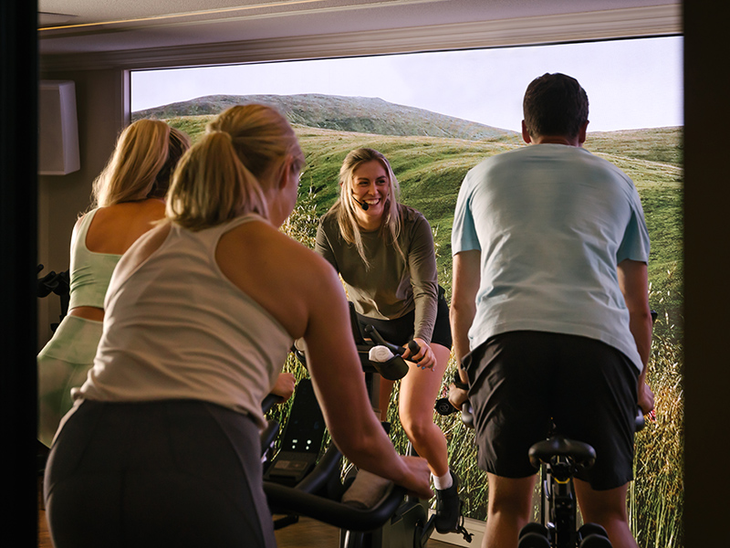 Four people take part in a stationary bike ride class in front of an interactive screen