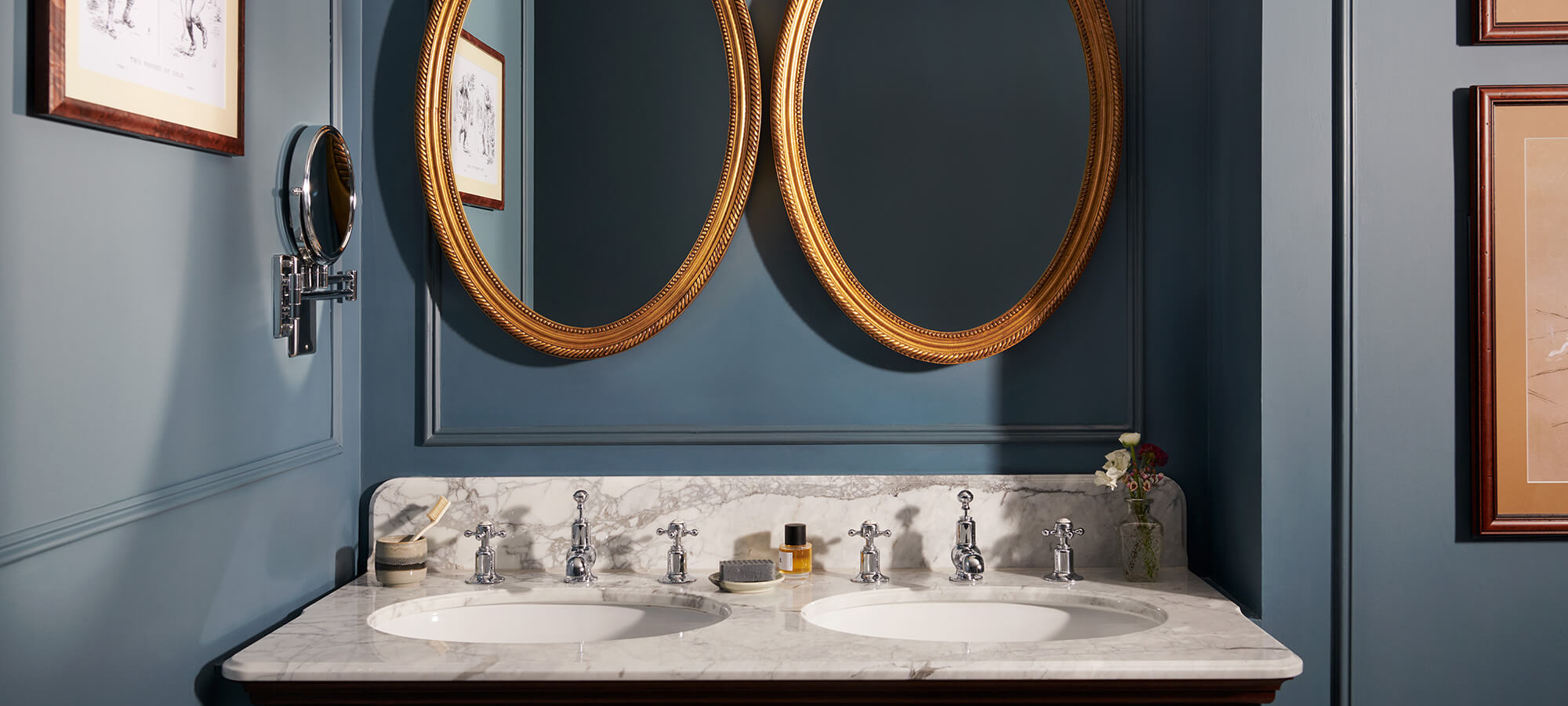 Zwei ovale goldene Spiegel über einem Doppelwaschbecken in einem marineblau gestrichenen Badezimmer in einem Estate-Zimmer des Haupthauses