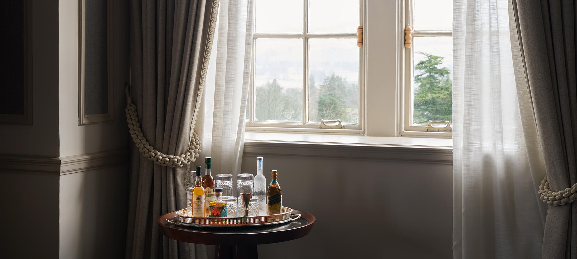 A small table with premium spirits and crystal glassware sits in front of a large window in a one bedroom whisky suite