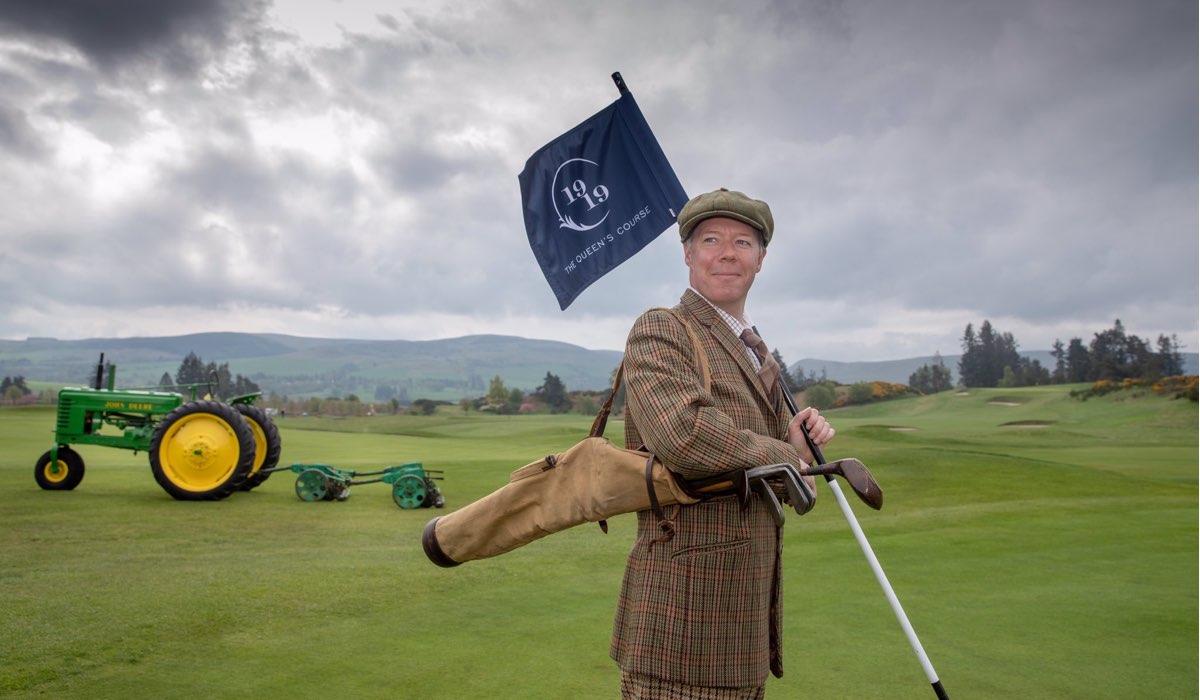 A man dressed in vintage golfing attire with a vintage tractor hold s a flag to commemorate the 100th anniversary of the King's and Queen's courses
