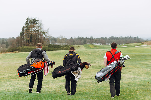 three golfers walk side by side with bags on their backs down a misty fairway