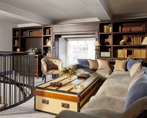 A large chest with glasses of whisky on top, sits in front of a large corner sofa surrounded by book cases
