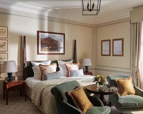 A large, brightly lit Estate bedroom, with paintings on the walls and two green tub chairs at the foot of the bed