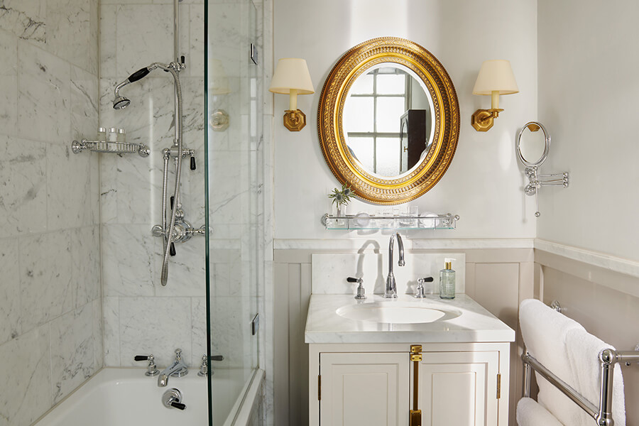 A marble shower above a large bath next to a single sink with gold mirror in a Country bedroom