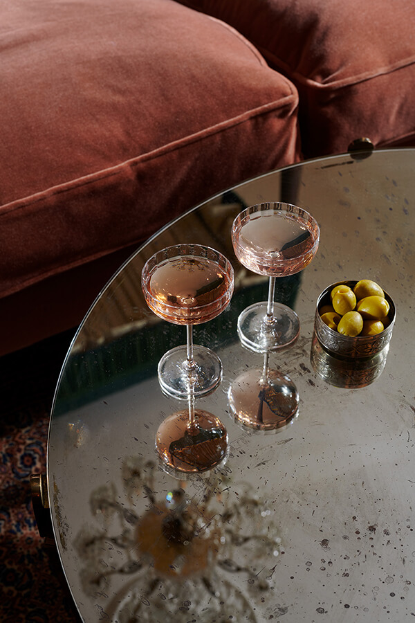 Two glasses of rose champagne and a ramekin of olives sit on a glass table next to a red sofa