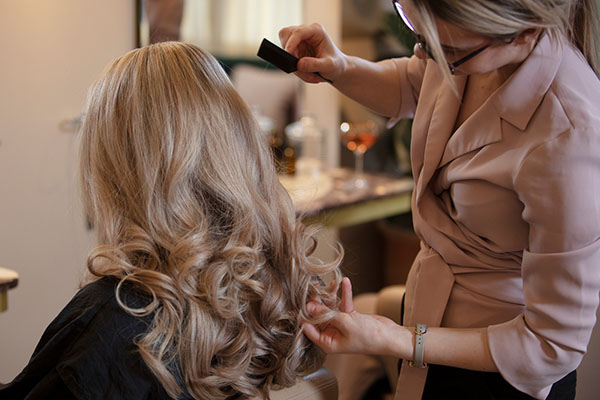 A stylist wearing a pink blouse curls a guest's blonde hair