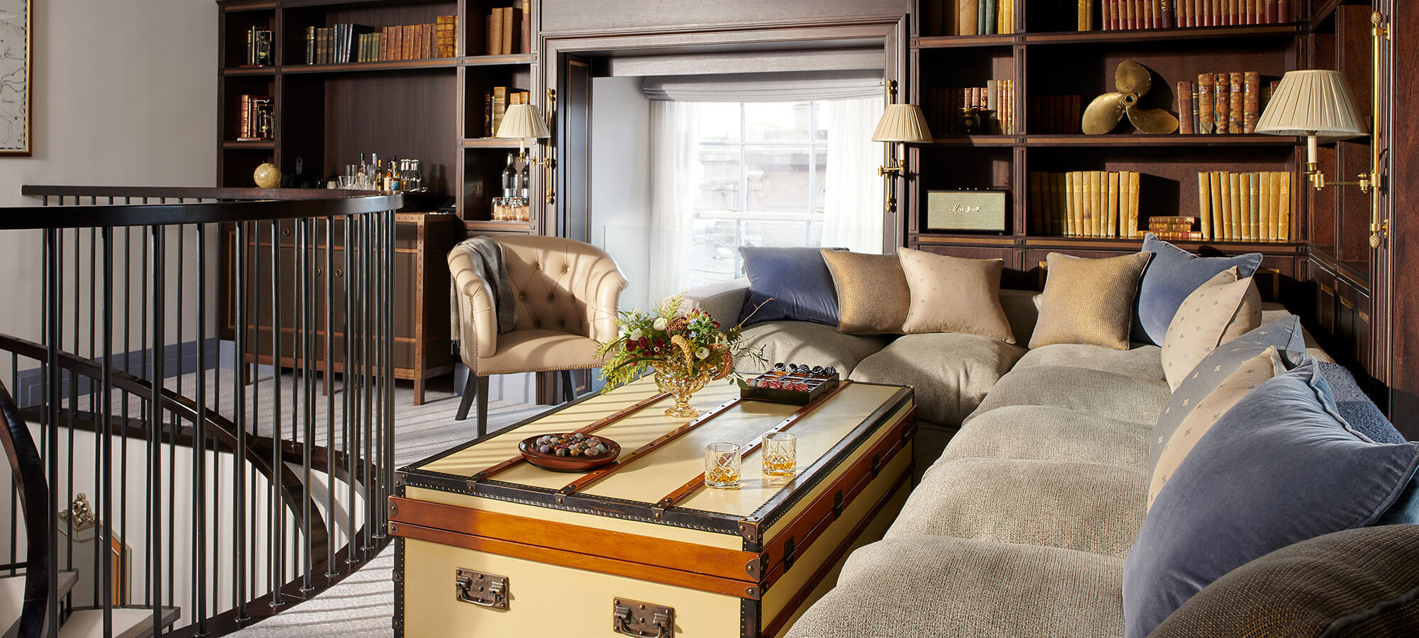 A large corner sofa covered in blue and grey cushions surrounds a chest ottoman at the top of a spiral staircase in the Tower Suite
