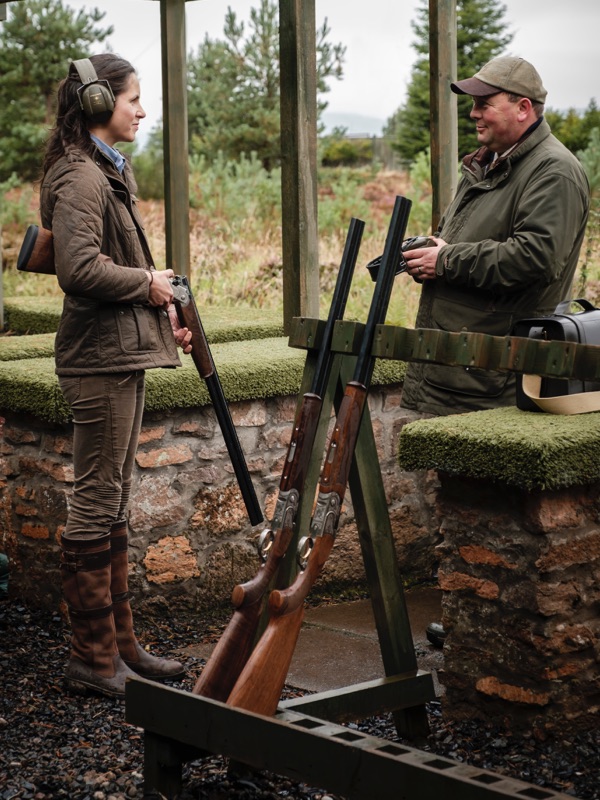 A woman speaks to a shooting instructor on a shooting range with a rack of shotguns next to them
