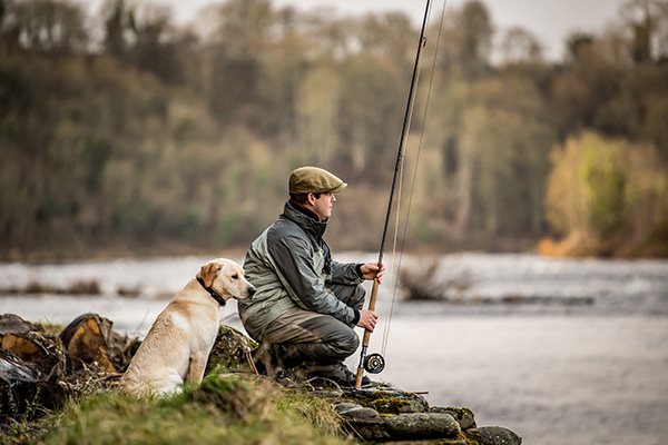 A fisherman with fly rod in hand and a Labrador sit on the banks of the River Tay
