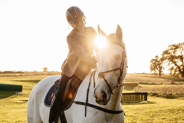 A rider pats her horse as the sun shines directly into the camera at sunset