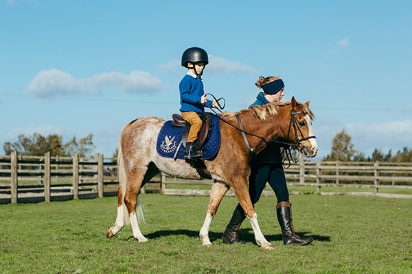 Un jeune cavalier portant un pull bleu assis sur un poney dirigé par un moniteur d'équitation.