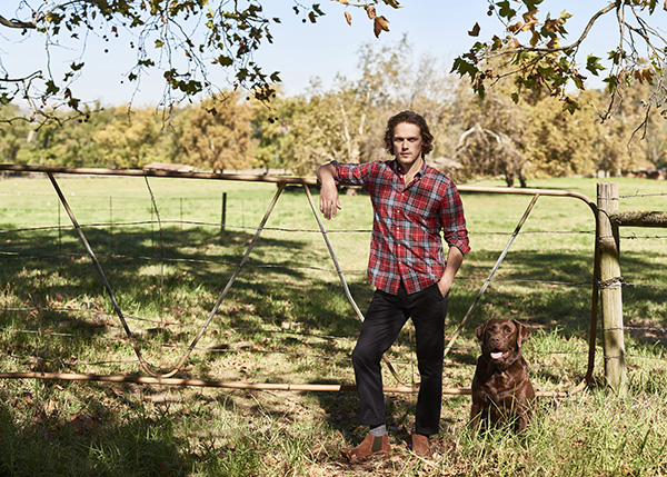Sam Sam Heaughan dressed in checked shirt kleaning on a fence with a Labrador
