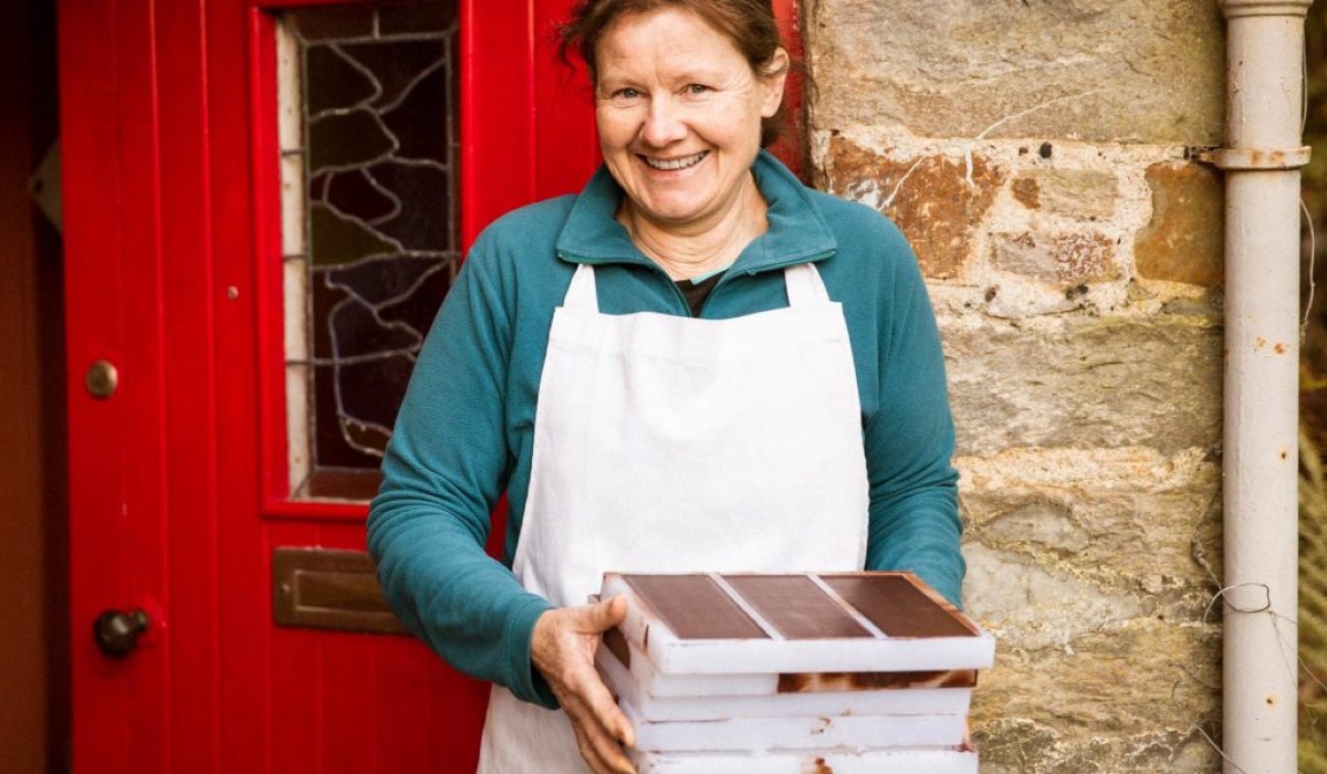 A woman stands with bars of chocolate outside a stone house with a red door.