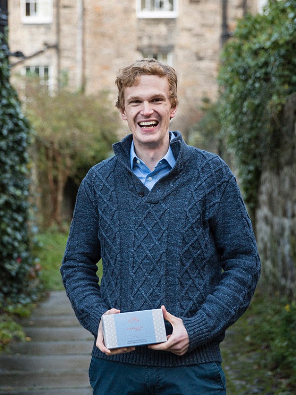 A smiling man with a blue jumper looks at the camera with a box of shortbread in his hands
