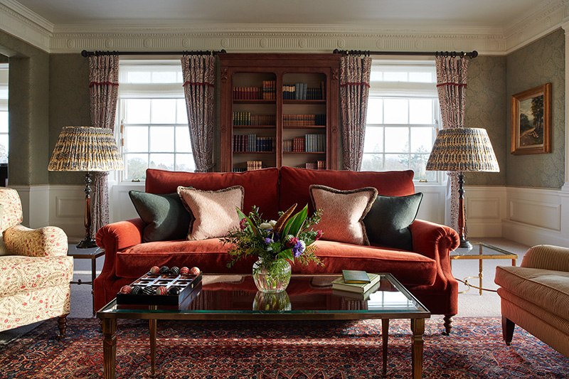 A large red sofa sits behind a glass table tin fresh flowers and in front of a book case filled with old books