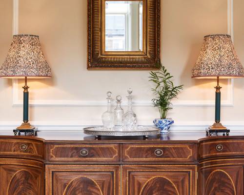 Crystal decanters on a silver tray with large lamp on top of an ornate side table in the assembly room