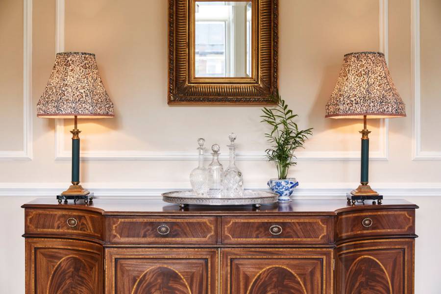 Crystal decanters on a silver tray with large lamp on top of an ornate side table in the assembly room