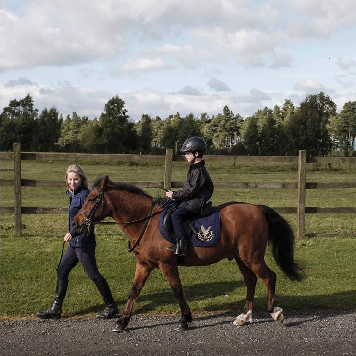 A riding instructor leads a young boy on a pony past a fence