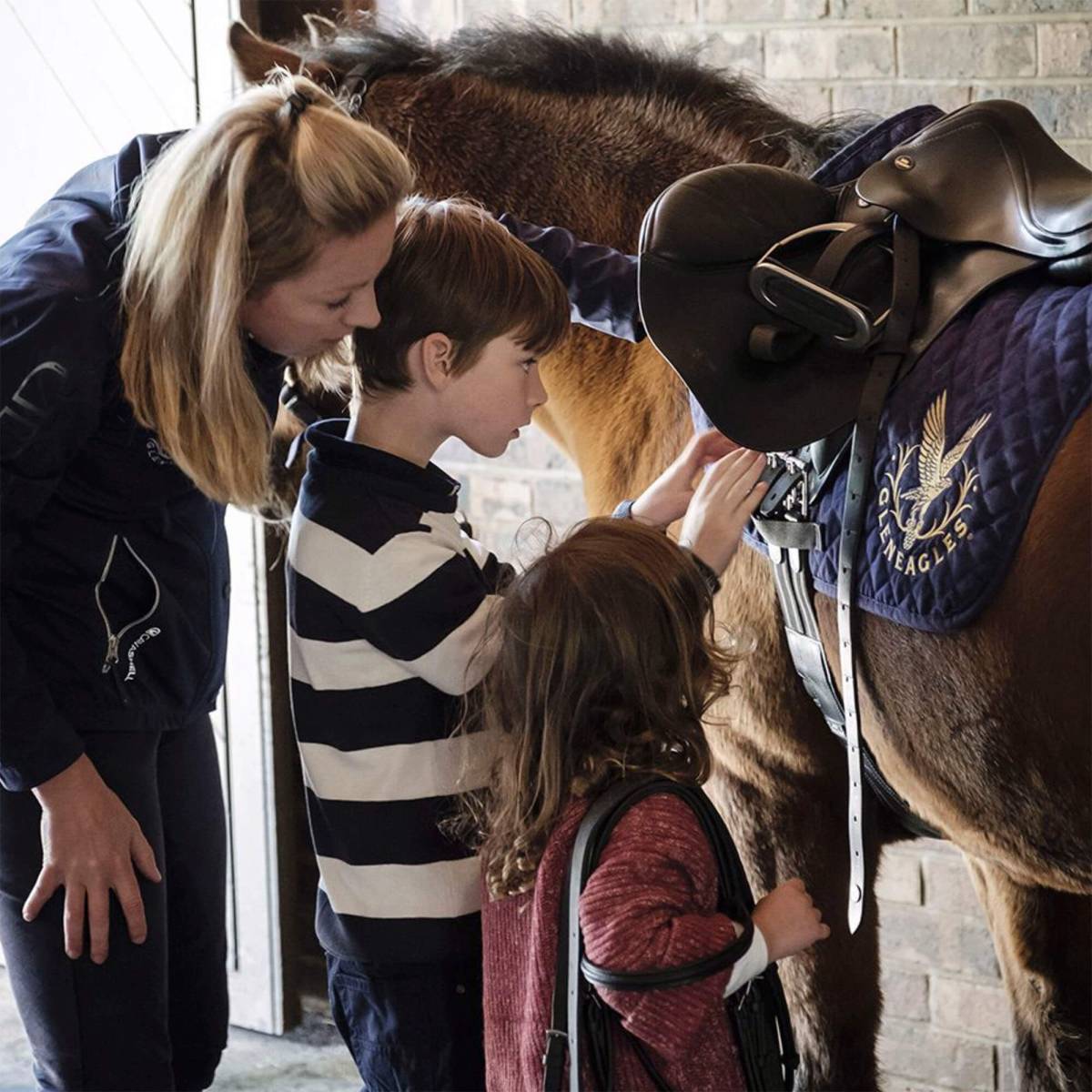 A young girl and boy are shown how to tack up a pony by a riding instructor