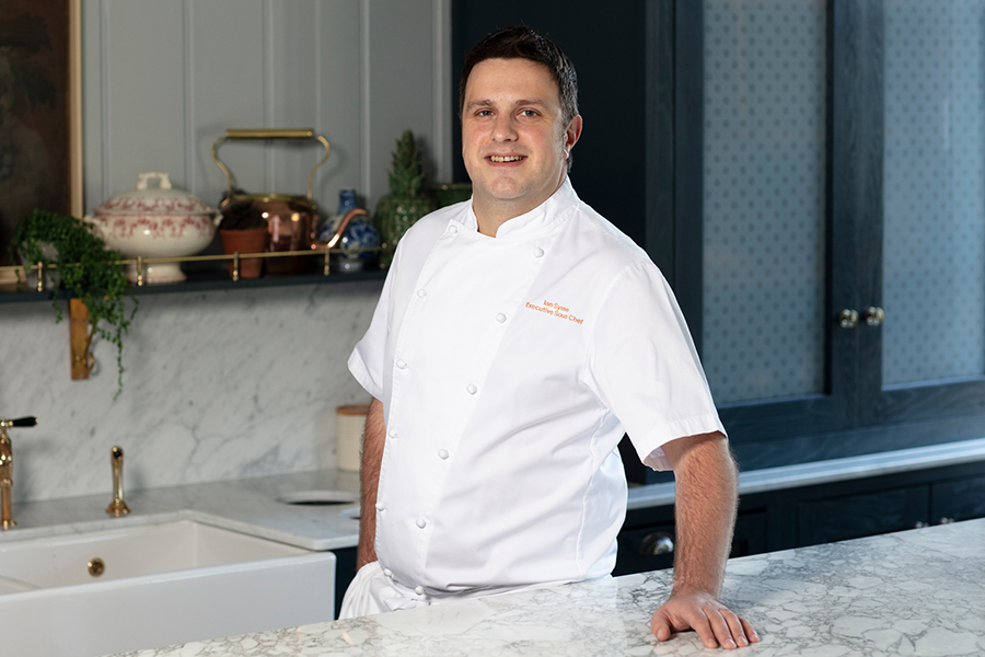 Chef Ian Syme stands with a hand on a marble worktop, smiling at the camera at Gleneagles
