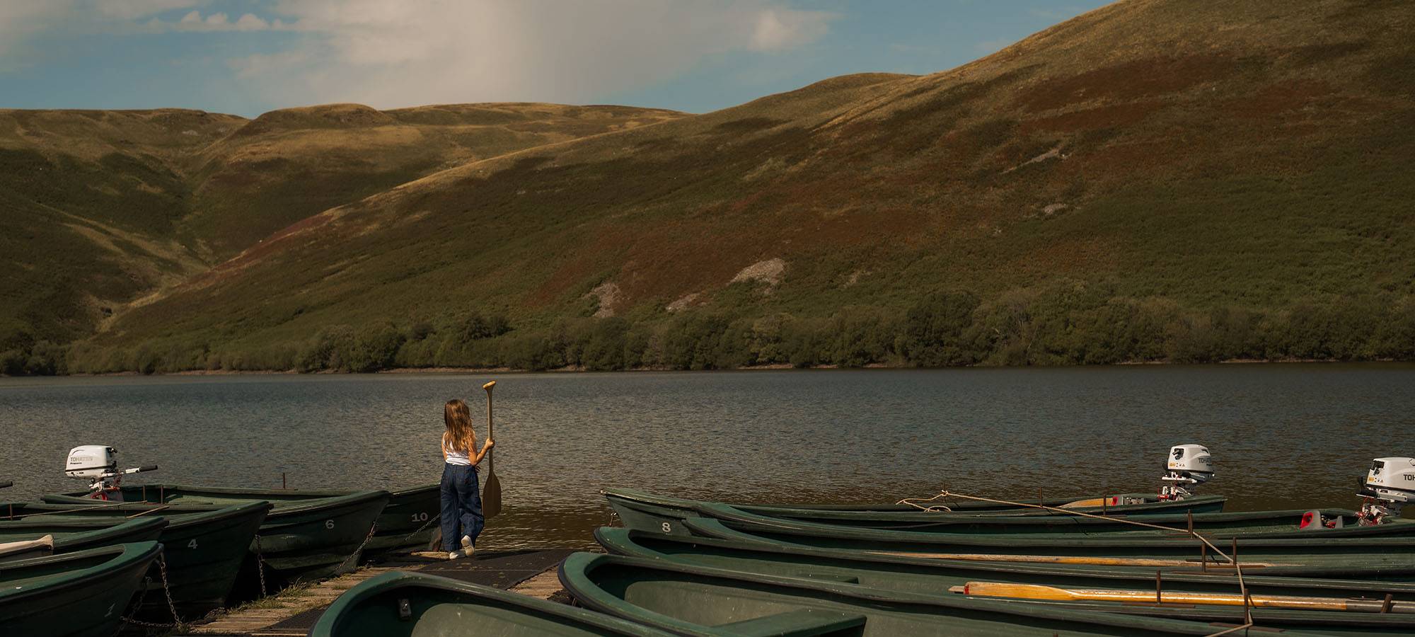 Landscape displays Ochil Hills and body of water, with a young girl standing on the boardwalk looking out