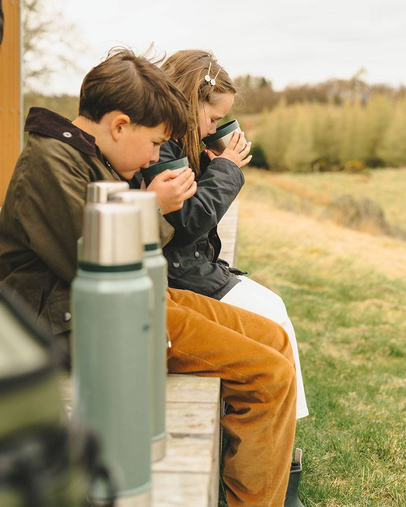 Young girl and young boy wearing outdoor jackets, drinking water from a flask.